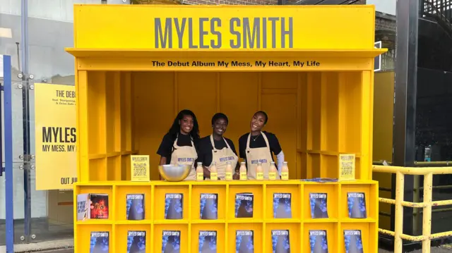 Three women smile inside a yellow stall with bottled coffees displayed on top and blue magazines with "Myles Smith" displayed on shelves along the front.