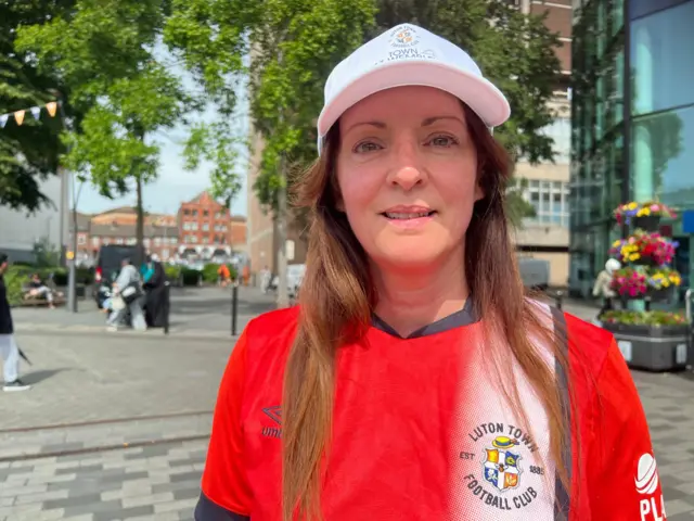 A woman wearing a Luton Town shirt and matching hat smiling as she stands in St George's Square