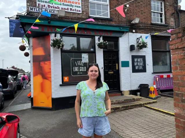 A woman wearing a green top and denim shorts is stood outside a pub on a road named "Myles Place".