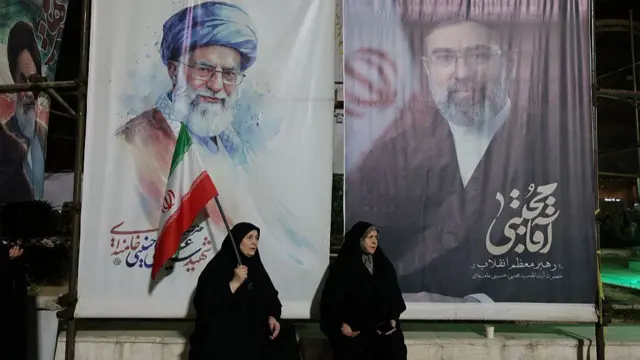 two women sitting in front of large posters with the faces of Ali Khamenei and his son Mojtaba Khamenei
