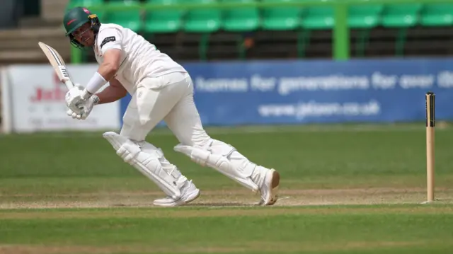 Lewis Hill, wearing a green helmet and white gloves while holding a cricket bat, looks over his shoulder after playing a shot while batting for Leicestershire