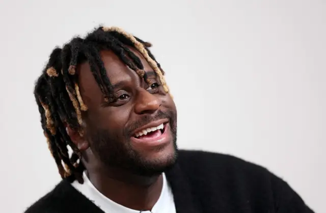 Myles Smith dressed in a black jumper and smiling against a blank white background
