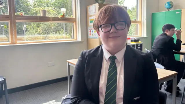 School pupil Alex wearing uniform sits in classroom setting and smiles at camera