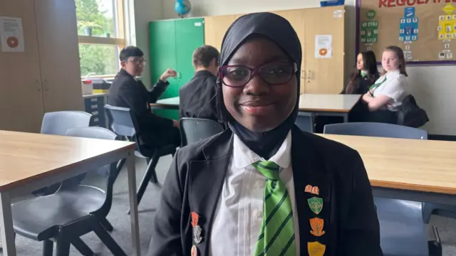 School pupil Aliyyah wearing uniform sits in classroom setting and smiles at camera