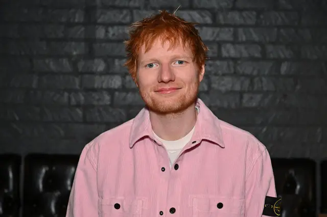 Ed Sheeran standing against a brick wall which has been painted black. Sheeran is smiling and wearing a pink jacket.