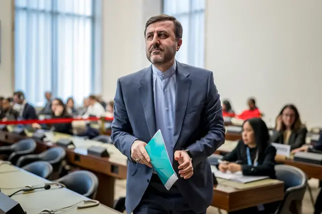 Iran's Deputy Foreign Minister Kazeem Gharibabadi standing up and walking around some desks and chairs holding a file