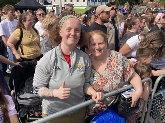 Two smiling women are stood at the barrier of the Myles gig. One is wearing a grey quater-zip and holding her thumb up.