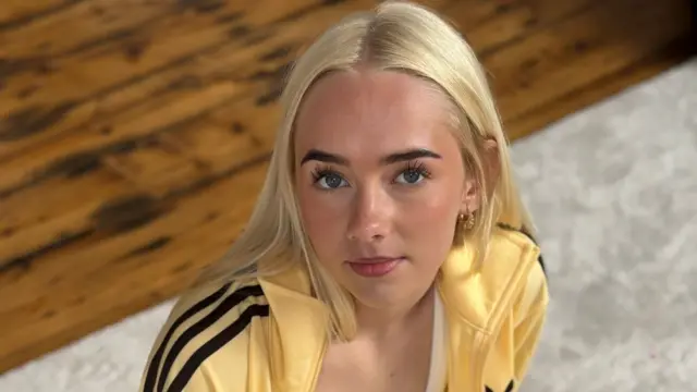 A blonde girl wearing a yellow tracksuit looks at the camera