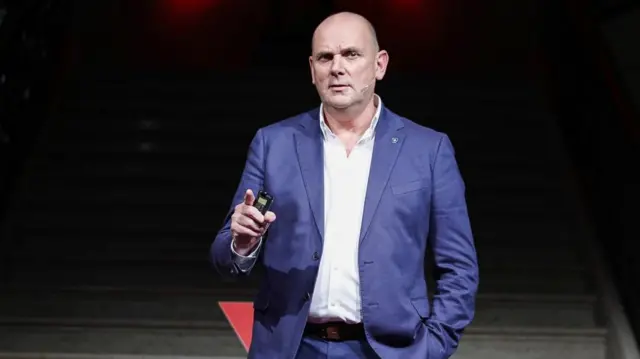 Jim Gamble, an older white man in a blue suit and white shirt speaking on stage.
