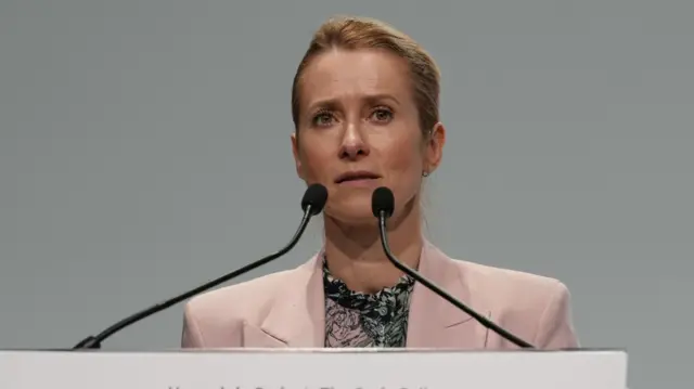 A woman, wearing a pink blazer, speaking into two microphones.