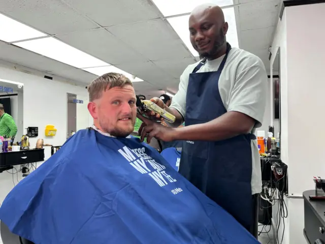 A man with short hair and a beard is getting his haircut by a barber holding clippers