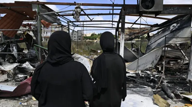 two women standing in front of a collapsed and destroyed building