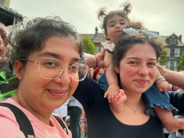 Two women smile with a young girl sat on the shoulders of one of them.