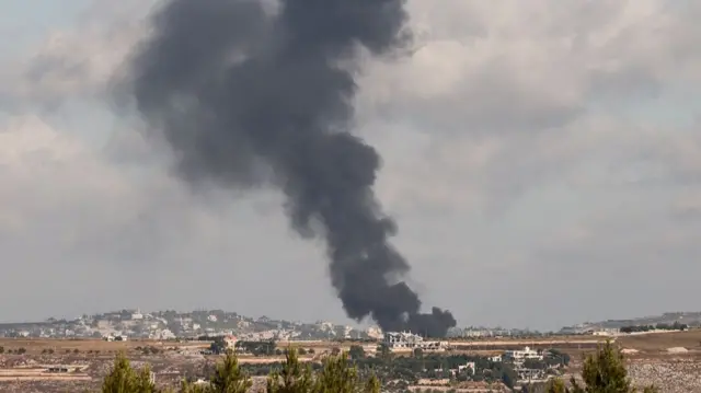 A photo of a wide landscape shows a dark cloud of smoke billowing from an inhabited area on the landscape
