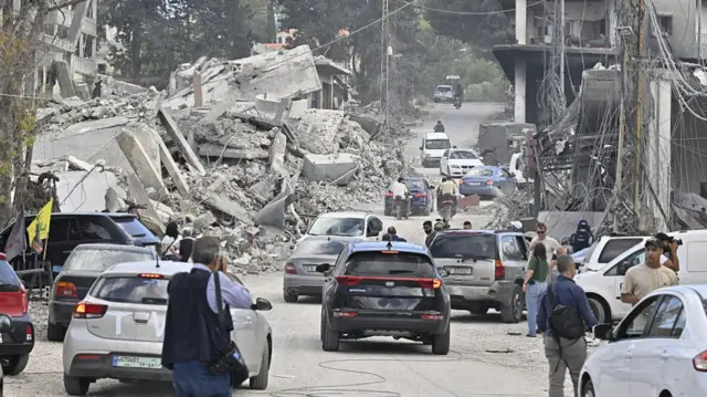 cars driving down a street and people standing around in the midst of massive piles of rubble where buildings used to stand