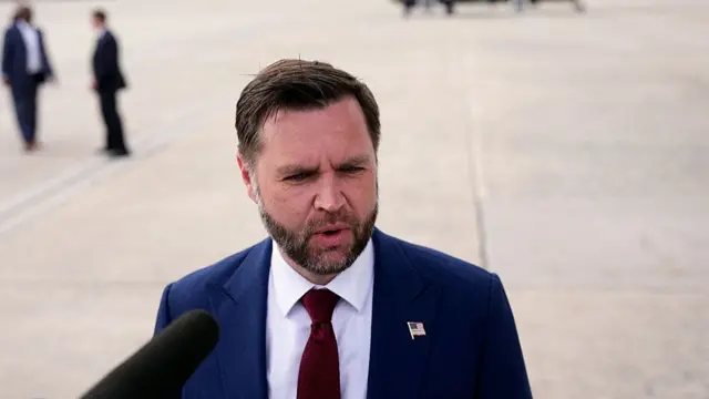 US Vice President JD Vance speaking to the press on an airport tarmac, two people to his left in the blurred background