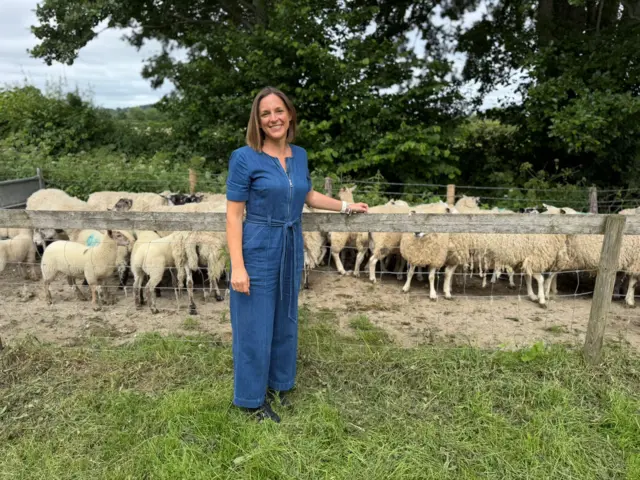A woman wearing a denim jumpsuit stands in front of a wooden fence which has sheep behind.