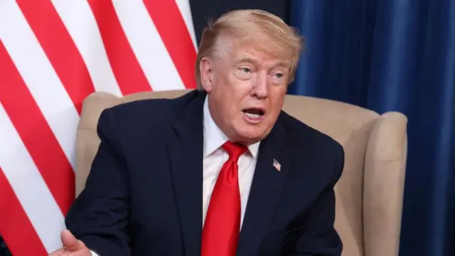 Donald Trump in a blue suit and red tie speaking while sat in a chair.