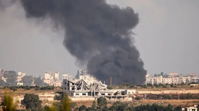 A photo of a black cloud of smoke billowing from a destroyed area in Lebanon. We can see debris from buildings in the foreground and more buildings in the background