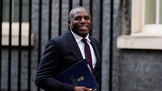 David Lammy walks up Downing Street, he wears a suit and holds a blue leather bound folder.