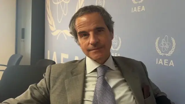 Rafael Grossi is pictured seated in a chair, wearing a grey jacket, white shirt and spotted blue tie. A blue wall behind him is adorned with the 'IAEA' logo.