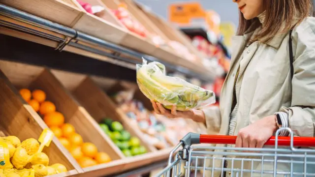 A woman selecting bananas from a supermarket produce aisle with a shopping cart.