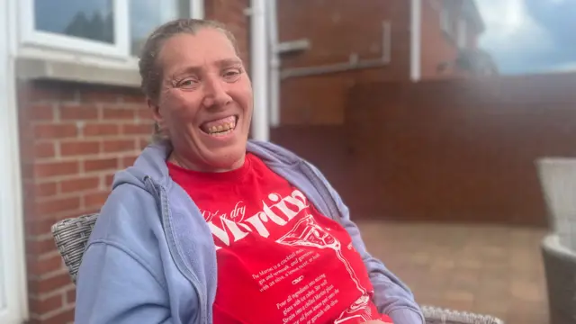 Laura is looking toward the camera and smiling. She is dressed in a red t-shirt with white text and is wearing a grey zip hoody.