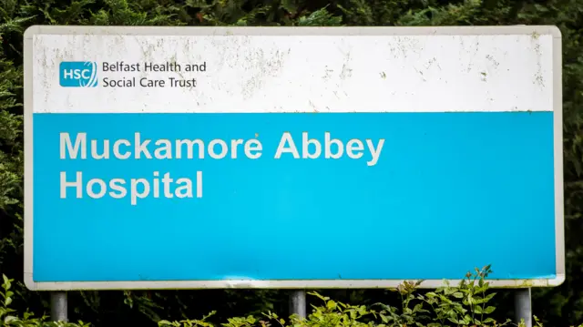The image shows a large blue and white sign. It reads Belfast Health and social Care Trust and below it is 'Muckamore Abbey Hospital'.