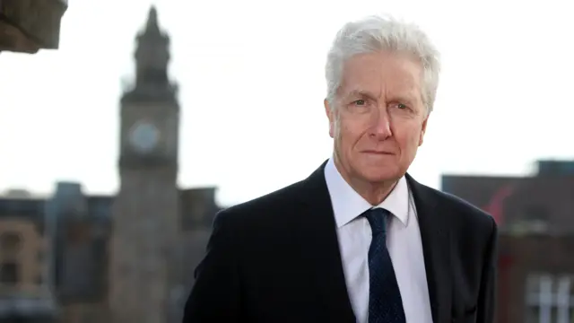 Tom Kark is looking into the camera. He has white hair and is dressed in a dark suit and tie with a white shirt.