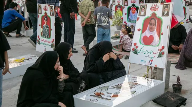 Women mourners dressed in black are shown gathered around memorial posters showing the faces of children who died in the strike.