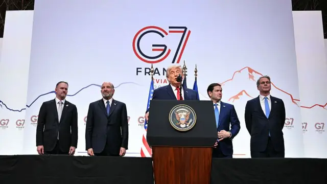 Donald Trump speaking at a podium at the G7 Summit with senior administration officials behind him including Marco Rubio