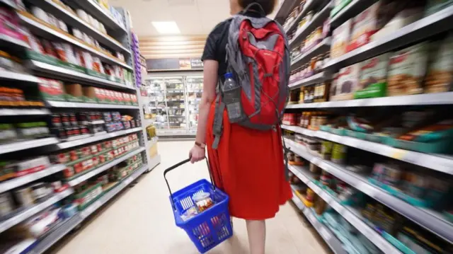 A woman in a red skirt and wearing a red backpack holds a blue shopping basket as she walks down a shopping aisle