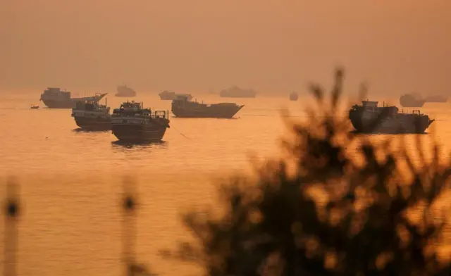 Around a dozen ships are shown in the water, with golden light suggesting it is around sunset.