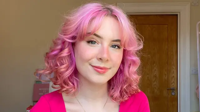 Milly Evans smiling in a photo in a bedroom, wearing a neon pink top