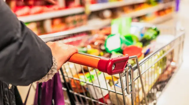 Person rolls a shopping trolley full of products down a supermarket aisle.