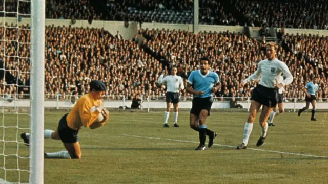 During the first game of the World Cup Soccer Championships at Wembley Stadium, July 11th, English goalie Gordon Banks (wearing yellow sweater) falls to his knees in effort to catch ball kicked by opposing Uruguayan player.
