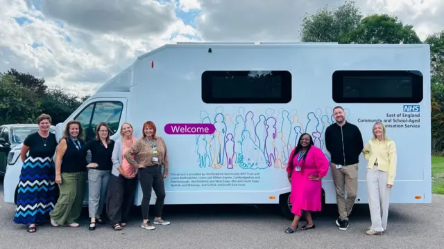 Eight people stand smiling, posing for a photo in front of a van that is used to deliver vaccinations for children. The people are wearing staff lanyards and the van is marked 'NHS East of England Community and School-Aged Immunisation Service'.