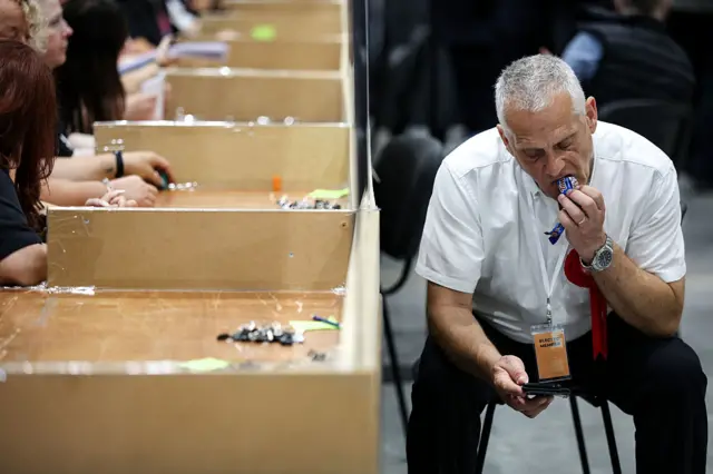 As people count votes, a man in a white shirt wearing a lanyard which says "election member" is mid-eating a chocolate bar as he checks his phone