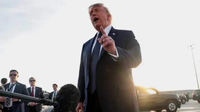 Donald Trump is pictured on a concrete tarmac speaking in front of a bank of microphones, wearing a suit and tie. He is gesturing with his left hand.