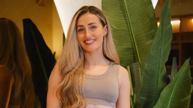 A woman with long blonde hair, blue eyes, smiling and wearing a gym outfit.