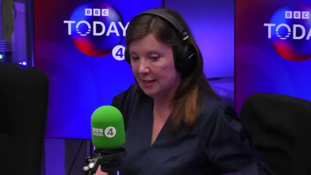 Rachel De Souza, who has straight brown hair and is wearing a dark blue blouse, sits in a radio studio wearing black headphones and speaking into a green microphone with the BBC Radio 4 logo printed on it.