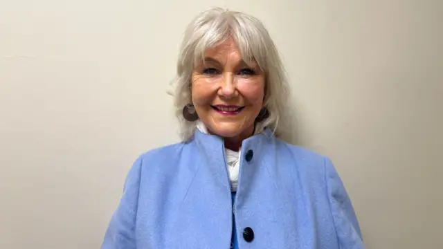 Margaret smiling at the camera. She is wearing hoop earrings and a pale blue jacket with large black buttons. She has long grey hair.