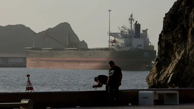 An oil tanker waiting in the Gulf of Oman. It's seen from the distance and in the foreground, two men stand on a pier.