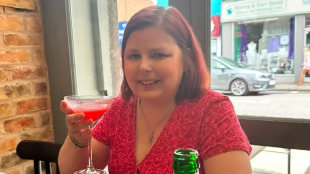 A young woman with pink, shoulder length hair is sat at a restaurant. She's holding up a drink and smiling to the camera