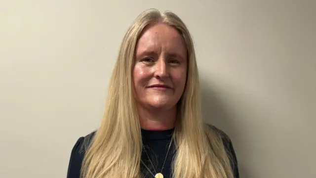 Mid-shot, woman with blonde hair, black top and gold necklace. Standing in front of a white wall