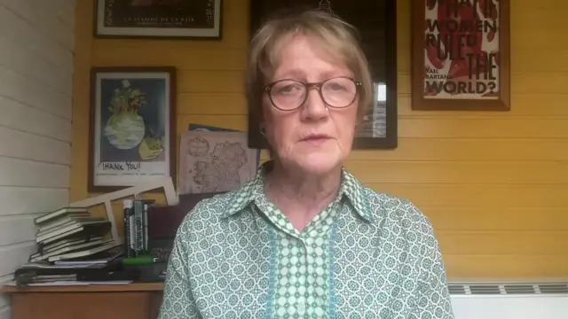 Patricia Lewis speaking to her webcam in her office. She's got round glasses on and a patterned, light green shirt