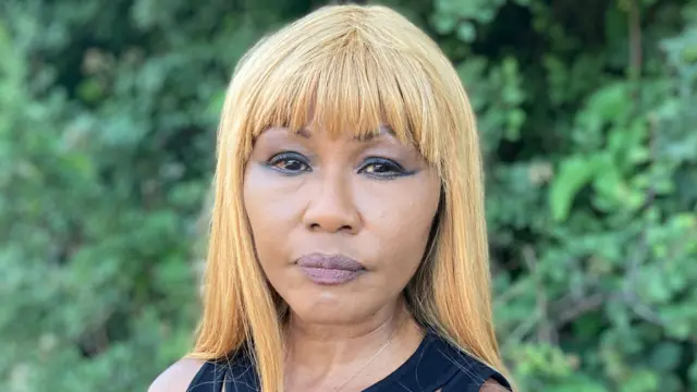 A woman with blonde hair looks at the camera. She is not smiling. She is wearing a black tank top and is standing in front of a green bush.