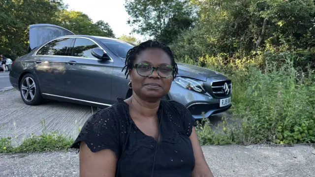 A woman sits on the ground outside, in front of a silver car. She has black hair and is wearing a black top.