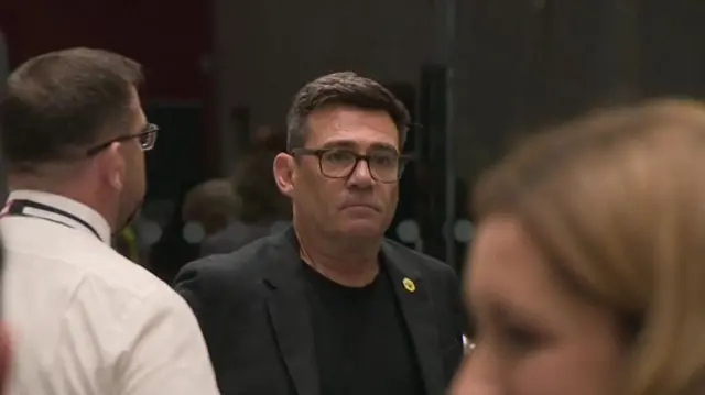 Andy Burnham is seen in a dark t-shirt and dark suit as he arrives at the Makerfield by-election count