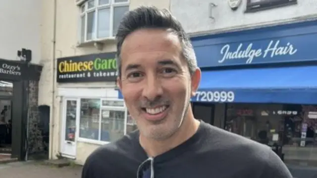 Tony Thakur, with a quiff stands in front of a hairdressers wearing a black tshirt and sunglasses.